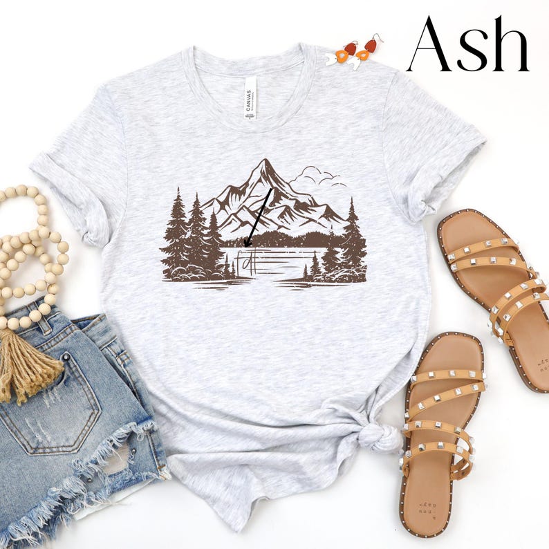 May include: A gray t-shirt with a brown mountain and lake graphic. The graphic features a mountain range with a lake in the foreground and a fishing rod in the air. The text "Ash" is in the upper right corner of the image.