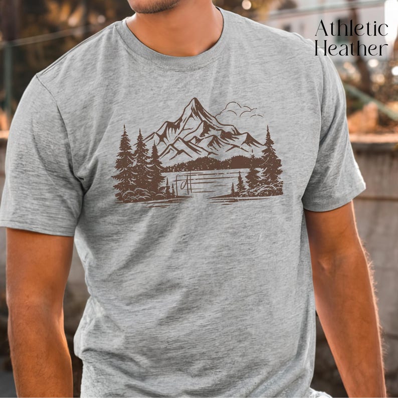 May include: A heather grey t-shirt with a brown mountain scene graphic. The graphic features a mountain range with a lake and trees.