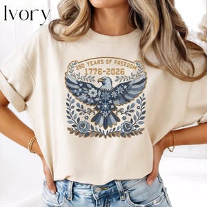 May include: Ivory-colored t-shirt featuring a patriotic design. The graphic includes an eagle with blue and white floral accents, the text "250 YEARS OF FREEDOM 1776-2026", and floral and star details. The shirt has short sleeves and a crew neck.