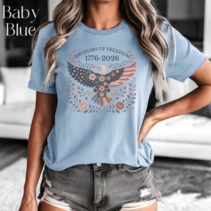 May include: Baby blue t-shirt featuring a graphic of an eagle with floral accents and the text "250 YEARS OF FREEDOM 1776-2026". The shirt has short sleeves and a crew neck.