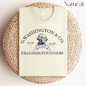May include: A cream-colored t-shirt with a graphic of George Washington and the text "G. WASHINGTON & CO. 1776 2026 250 YEARS OF LIBERTY FREEDOM FOUNDERS". The shirt is folded and placed on a woven, circular mat.