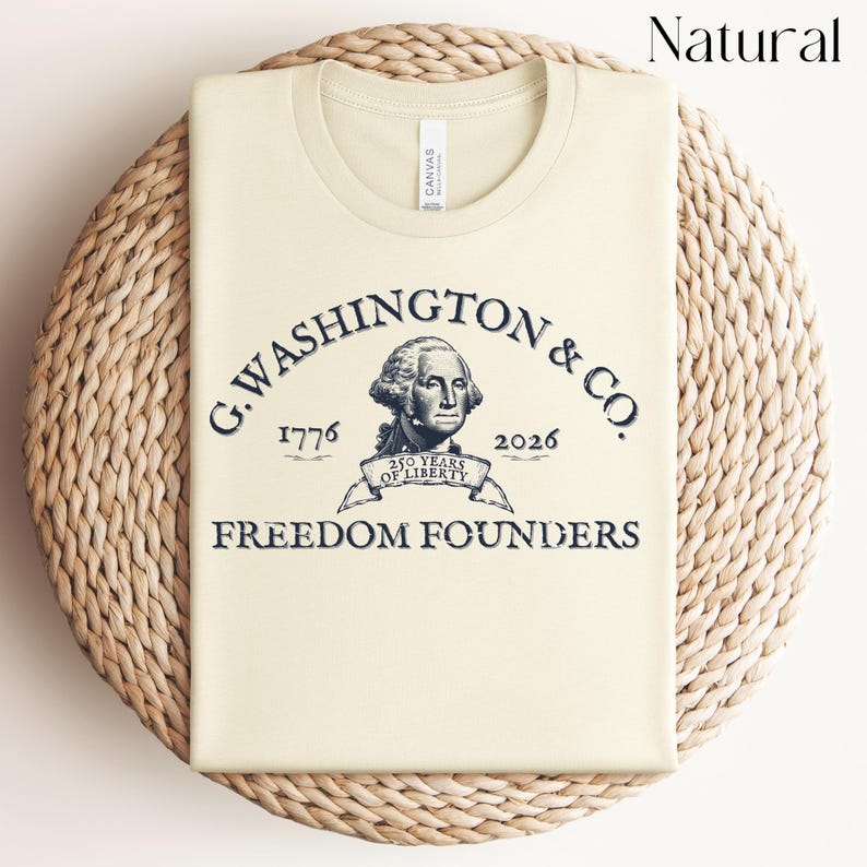 May include: A cream-colored t-shirt with a graphic of George Washington and the text "G. WASHINGTON & CO. 1776 2026 250 YEARS OF LIBERTY FREEDOM FOUNDERS". The shirt is folded and placed on a woven, circular mat.