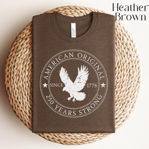 May include: Heather brown t-shirt with a white graphic of an eagle inside a circular design. The design includes the text "AMERICAN ORIGINAL SINCE 1776 250 YEARS STRONG". The shirt is folded and placed on a woven, circular mat.