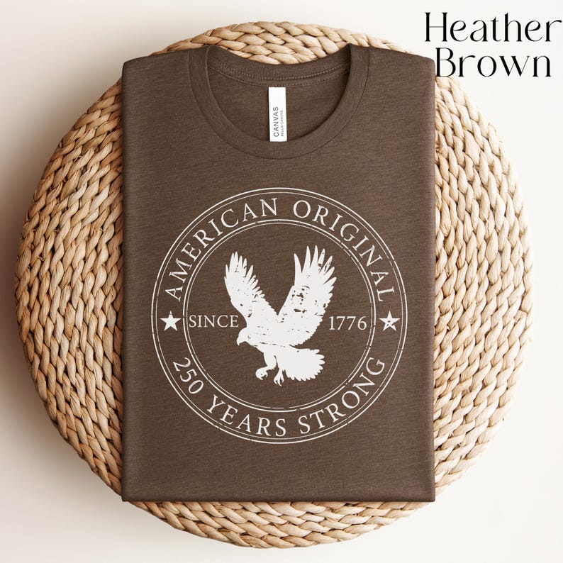 May include: Heather brown t-shirt with a white graphic of an eagle inside a circular design. The design includes the text "AMERICAN ORIGINAL SINCE 1776 250 YEARS STRONG". The shirt is folded and placed on a woven, circular mat.