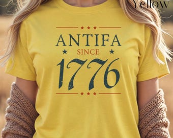 May include: Maize yellow t-shirt with the text "ANTIFA SINCE 1776" in navy blue. The text is arranged with stars and lines above and below the words. The shirt is short-sleeved.