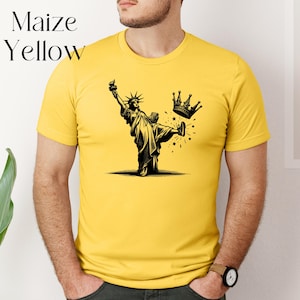 Może przedstawiać: Kukurydziana ż&oacute;łta koszulka z czarno-białą ilustracją Statuy Wolności kopiącej koronę. Tekst "Maize Yellow" znajduje się w lewym g&oacute;rnym rogu. Koszulka ma kr&oacute;tkie rękawy i okrągły dekolt.