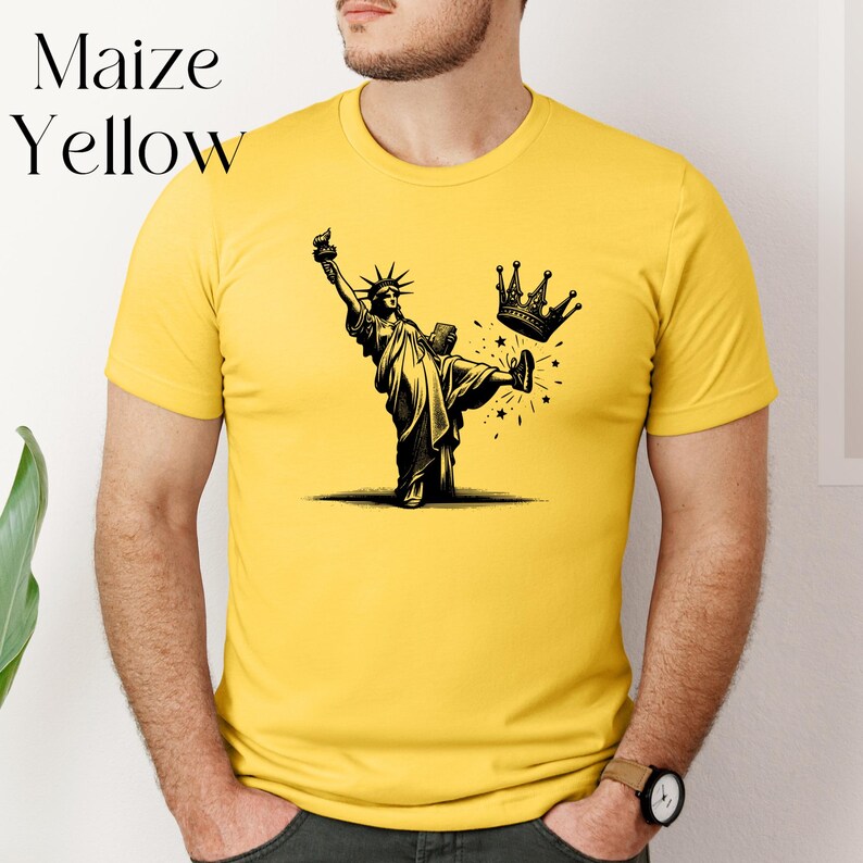 Może przedstawiać: Kukurydziana ż&oacute;łta koszulka z czarno-białą ilustracją Statuy Wolności kopiącej koronę. Tekst "Maize Yellow" znajduje się w lewym g&oacute;rnym rogu. Koszulka ma kr&oacute;tkie rękawy i okrągły dekolt.