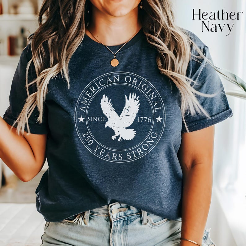 May include: Heather navy t-shirt with a white circular graphic featuring an eagle and the text "AMERICAN ORIGINAL SINCE 1776 250 YEARS STRONG". The shirt has rolled-up sleeves and is paired with blue jeans.
