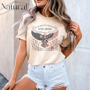 May include: A beige t-shirt featuring a patriotic design. The graphic includes an eagle with a floral design, the text "250 YEARS OF FREEDOM 1776-2026", and the word "Natural" at the top left.