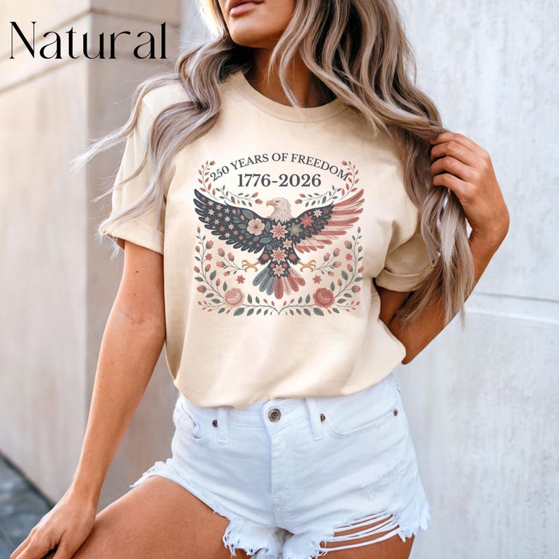 May include: A beige t-shirt featuring a patriotic design. The graphic includes an eagle with a floral design, the text "250 YEARS OF FREEDOM 1776-2026", and the word "Natural" at the top left.