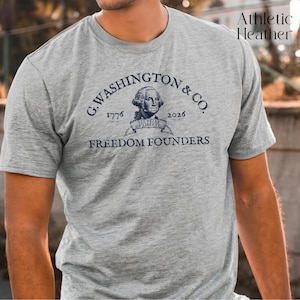 May include: A gray athletic heather t-shirt featuring the text "G. Washington & Co." with the years 1776 and 2026, and the words "Freedom Founders." The shirt has a graphic of a historical figure and the text "250 Years of Liberty."