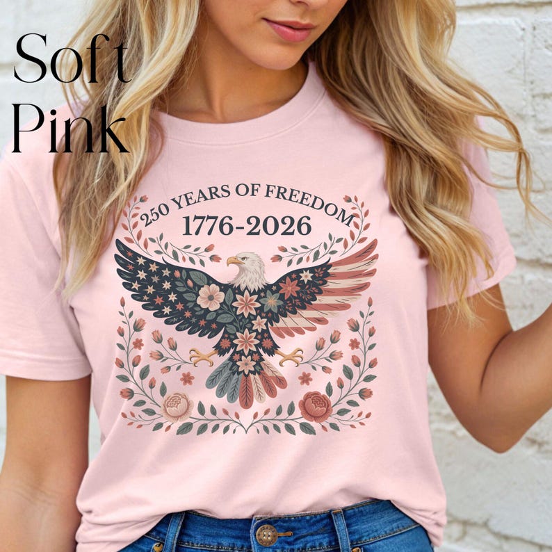 May include: Soft pink t-shirt featuring a patriotic design. The graphic includes an eagle with a floral pattern, stars, and the text "250 YEARS OF FREEDOM 1776-2026". The shirt is a light pink color.