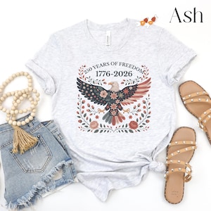 May include: Light gray t-shirt with a patriotic design featuring an eagle with floral accents and the text "250 YEARS OF FREEDOM 1776-2026". The shirt is paired with denim shorts and sandals, suggesting a casual, everyday style.