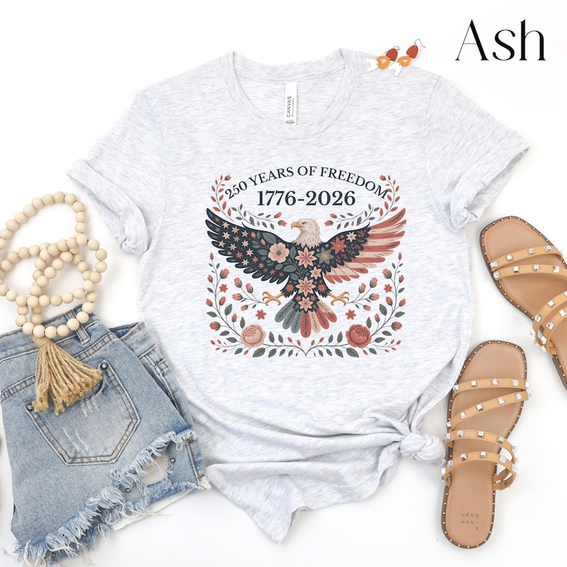 May include: Light gray t-shirt with a patriotic design featuring an eagle with floral accents and the text "250 YEARS OF FREEDOM 1776-2026". The shirt is paired with denim shorts and sandals, suggesting a casual, everyday style.