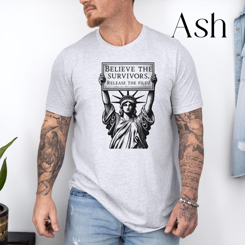 May include: Light grey t-shirt with a black and white graphic of the Statue of Liberty holding a sign. The sign reads "BELIEVE THE SURVIVORS. RELEASE THE FILES!" The shirt is a light ash colour.