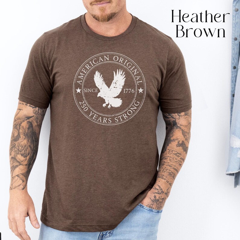 May include: Heather brown t-shirt with a circular graphic featuring a white eagle and the text "AMERICAN ORIGINAL SINCE 1776 250 YEARS STRONG". The shirt is a solid color and has short sleeves.