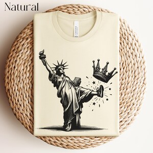 Może przedstawiać: Kremowa koszulka z czarno-białą ilustracją Statuy Wolności kopiącej koronę. Tekst "Natural" znajduje się w lewym g&oacute;rnym rogu. Koszulka ma okrągły dekolt i metkę "Canvas".