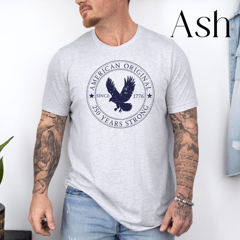 May include: Ash-colored t-shirt featuring a navy blue circular graphic with an eagle and the text "AMERICAN ORIGINAL SINCE 1776 250 YEARS STRONG". The shirt is a light gray color.