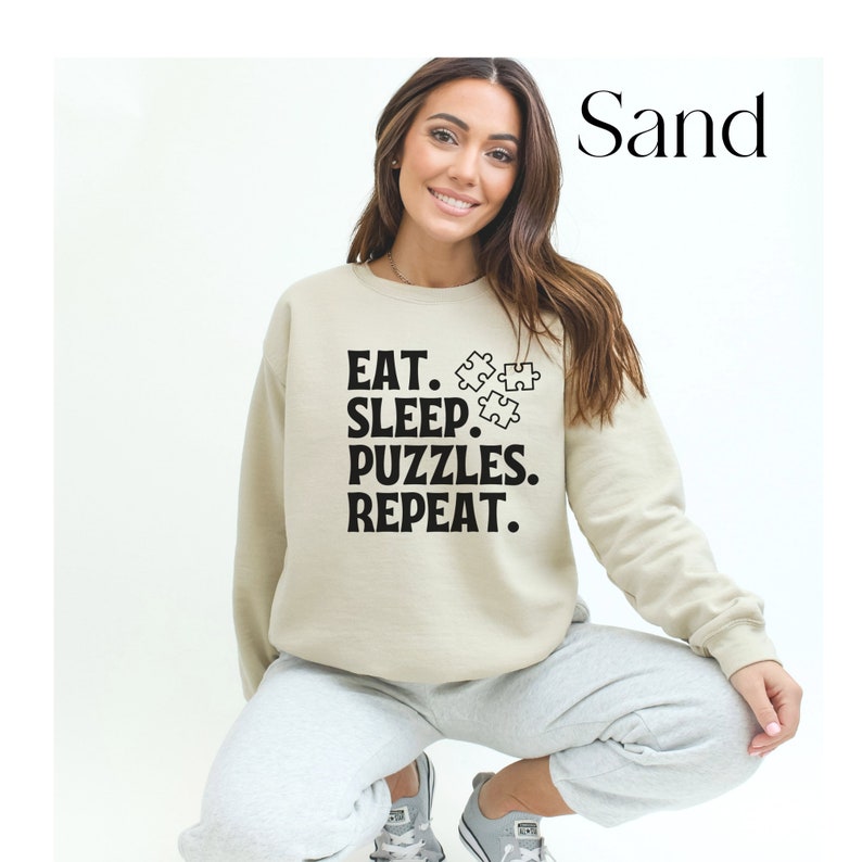 May include: A beige sweatshirt with the text "EAT. SLEEP. PUZZLES. REPEAT." in black. The text is accompanied by a graphic of three puzzle pieces.