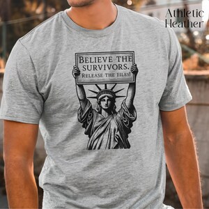 May include: Athletic heather grey t-shirt with a black and white graphic of the Statue of Liberty holding a sign. The sign reads "BELIEVE THE SURVIVORS. RELEASE THE FILES!" The shirt is short-sleeved.