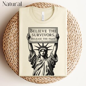 May include: A cream-coloured t-shirt with a black and white graphic of the Statue of Liberty holding a sign that reads "BELIEVE THE SURVIVORS. RELEASE THE FILES!" The shirt has a crew neck and a "CANVAS" label.
