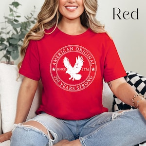 May include: A red t-shirt with a white circular graphic featuring an eagle and the text "AMERICAN ORIGINAL SINCE 1776 250 YEARS STRONG." The word "Red" is in the upper right corner. The shirt is short-sleeved.