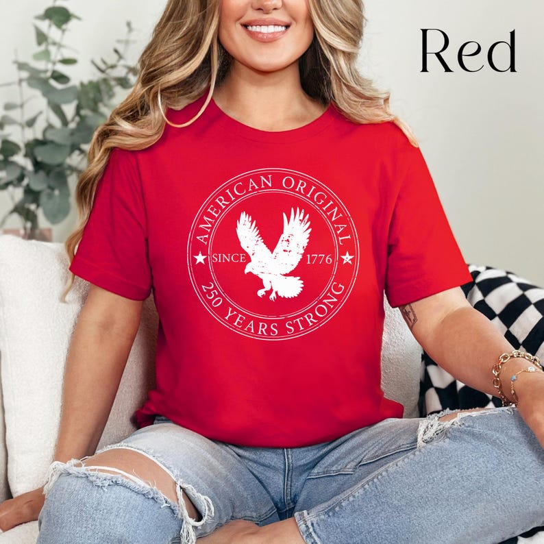 May include: A red t-shirt with a white circular graphic featuring an eagle and the text "AMERICAN ORIGINAL SINCE 1776 250 YEARS STRONG." The word "Red" is in the upper right corner. The shirt is short-sleeved.