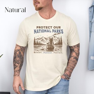 May include: A beige t-shirt with a brown bear and cub illustration. The text "Protect Our National Parks" is printed in blue and brown. The text "Resist. Persist. Prevail." is printed in brown below the illustration.