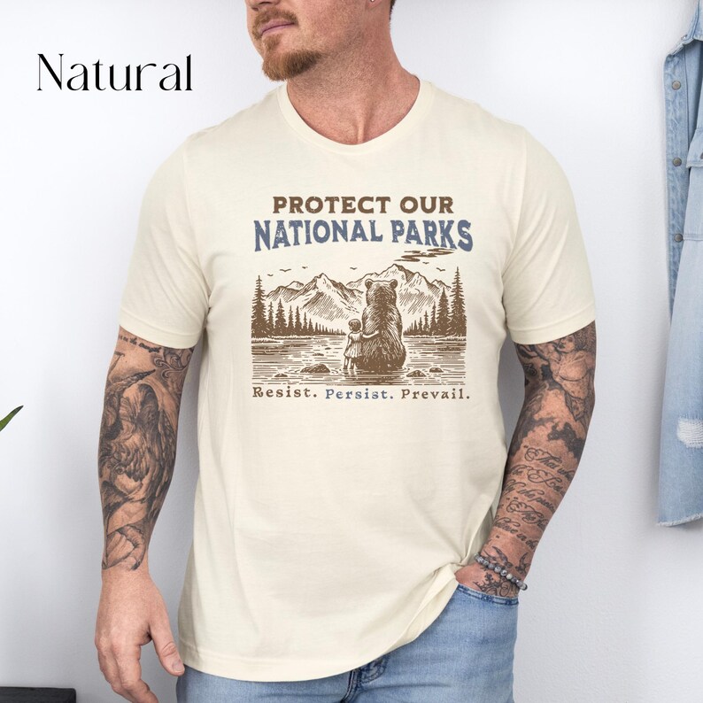 May include: A beige t-shirt with a brown bear and cub illustration. The text "Protect Our National Parks" is printed in blue and brown. The text "Resist. Persist. Prevail." is printed in brown below the illustration.