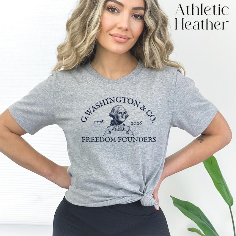 May include: Athletic Heather gray t-shirt with a crew neck. The shirt features a navy blue graphic of George Washington and the text "G. Washington & Co. 1776-2026 Freedom Founders". The shirt is made of a soft material.
