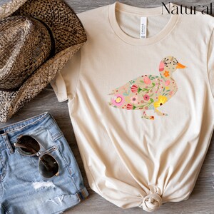 Peut inclure: Un t-shirt beige avec un motif de canard floral. Le canard est d&eacute;limit&eacute; par un contour noir et rempli de fleurs color&eacute;es. Le t-shirt est fait d'un tissu doux et confortable, parfait pour tous les jours.