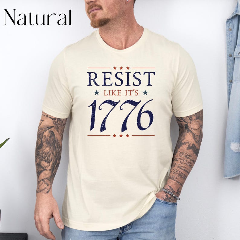Pode incluir: Camiseta creme com a frase "RESIST LIKE IT'S 1776" em azul marinho, com detalhes em vermelho. O texto est&aacute; centralizado na frente. A camiseta tem mangas curtas e gola redonda.