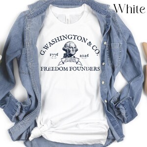 May include: White t-shirt with a navy blue graphic featuring an illustration of George Washington and the text "G. Washington & Co. 1776-2026 Freedom Founders". The shirt is layered under a light blue denim jacket.