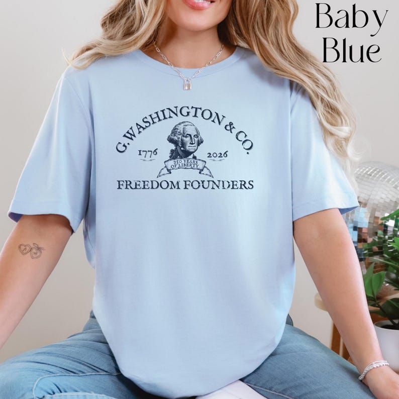 May include: Baby blue t-shirt featuring the text "G. Washington & Co. Freedom Founders" with a portrait of George Washington. The shirt also includes the years 1776 and 2026. The shirt is a light blue color.