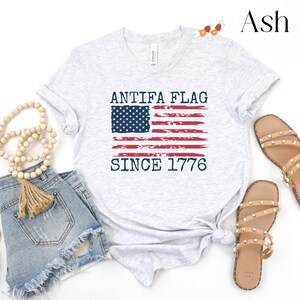 May include: Ash-colored t-shirt with the text "ANTIFA FLAG SINCE 1776" in navy blue. The design features a distressed American flag. The shirt is displayed with denim shorts and sandals.
