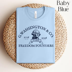 May include: Baby blue t-shirt with a graphic of George Washington and the text "G. WASHINGTON & CO. 1776-2026 FREEDOM FOUNDERS". The shirt is folded on a woven, circular surface.