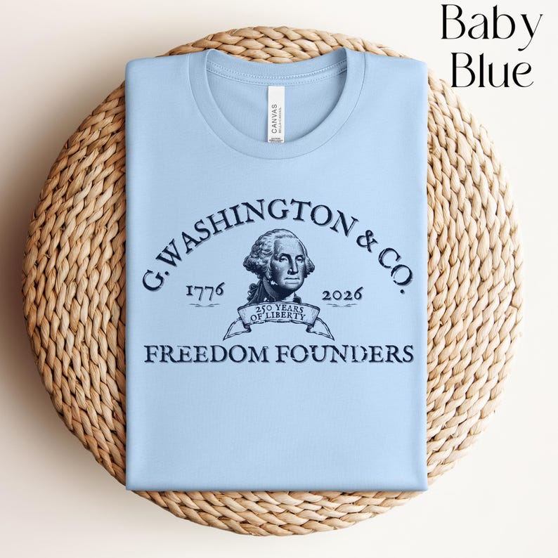 May include: Baby blue t-shirt with a graphic of George Washington and the text "G. WASHINGTON & CO. 1776-2026 FREEDOM FOUNDERS". The shirt is folded on a woven, circular surface.
