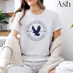 May include: Ash-colored t-shirt featuring a navy blue circular graphic with an eagle and the text "AMERICAN ORIGINAL SINCE 1776 250 YEARS STRONG". The shirt is short-sleeved and has a crew neck.