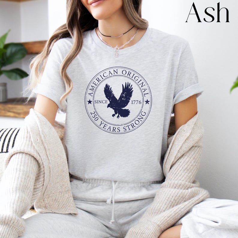 May include: Ash-colored t-shirt featuring a navy blue circular graphic with an eagle and the text "AMERICAN ORIGINAL SINCE 1776 250 YEARS STRONG". The shirt is short-sleeved and has a crew neck.