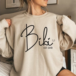 May include: Beige sweatshirt with the name "Bibi" in a black script font. Below the name is "EST 2019" and a small heart. The long-sleeved top is a neutral color.