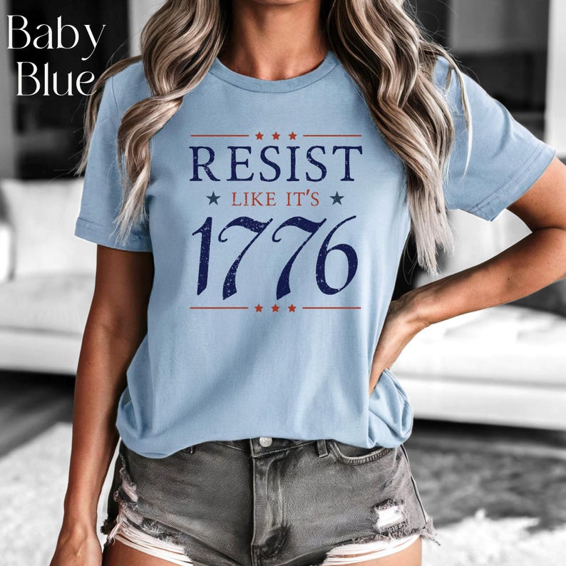 Pode incluir: T-shirt azul claro com a frase "RESIST LIKE IT'S 1776" em azul marinho, emoldurada por estrelas e linhas vermelhas. O texto "Baby Blue" &eacute; vis&iacute;vel no canto superior esquerdo. Manga curta.