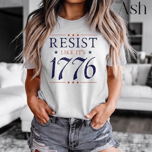 Pode incluir: T-shirt cinza claro com o texto "RESIST LIKE IT'S 1776" em azul marinho e vermelho. O design inclui estrelas e linhas acima e abaixo do texto. Uma pe&ccedil;a de roupa casual.