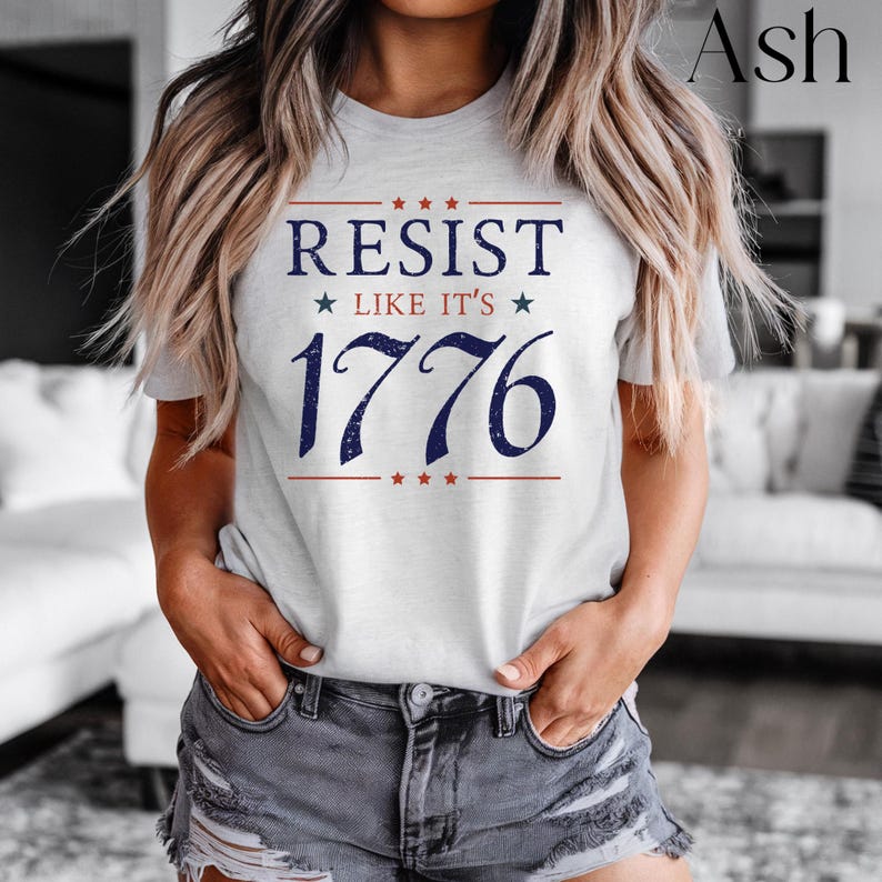 Pode incluir: T-shirt cinza claro com o texto "RESIST LIKE IT'S 1776" em azul marinho e vermelho. O design inclui estrelas e linhas acima e abaixo do texto. Uma pe&ccedil;a de roupa casual.