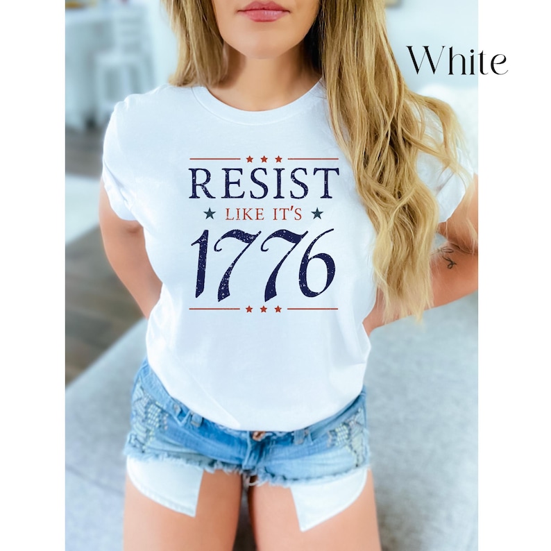 Pode incluir: T-shirt branca com o texto "RESIST LIKE IT'S 1776" em azul marinho e vermelho. A camisa tem gola redonda e mangas curtas. A palavra "White" est&aacute; escrita em uma fonte de script no canto superior direito.