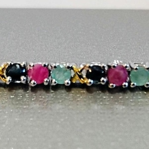May include: A delicate bracelet featuring a series of colorful gemstones. The bracelet alternates between red, green, and blue gemstones, set in a silver and gold-toned metal. The gemstones are round and evenly spaced.