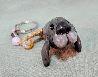 May include: A handmade manatee keychain. The manatee is gray with black details and small flipper-like appendages. The keychain includes a silver ring and decorative beads in pink, purple, and clear with gold flecks.
