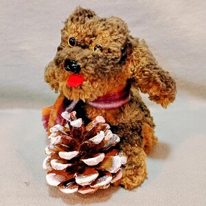 May include: A plush brown dog with a red tongue and black nose, wearing a scarf with orange, purple, and white stripes. A pine cone with white-painted tips rests in front of the dog. The dog has yellow eyes.