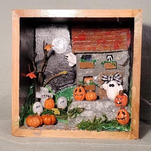 May include: A Halloween-themed shadow box with a miniature haunted house, pumpkins, ghosts, and a graveyard. The house has a red brick roof and a gray stone facade. The scene is set against a dark, starry background, with a full moon and a bare tree.
