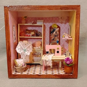 May include: A miniature diorama of a sewing room in a wooden box. The scene includes a pink sewing machine, a dress form with a white lace garment, and a small basket. Shelves and a cabinet hold sewing supplies.