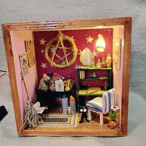 May include: Miniature witch's room diorama in a wooden box. The room features a broom, a rug, a black cat figurine, a chair, a shelf with bottles, and a pentagram on the wall. The walls are pink and red, with small lights illuminating the scene.